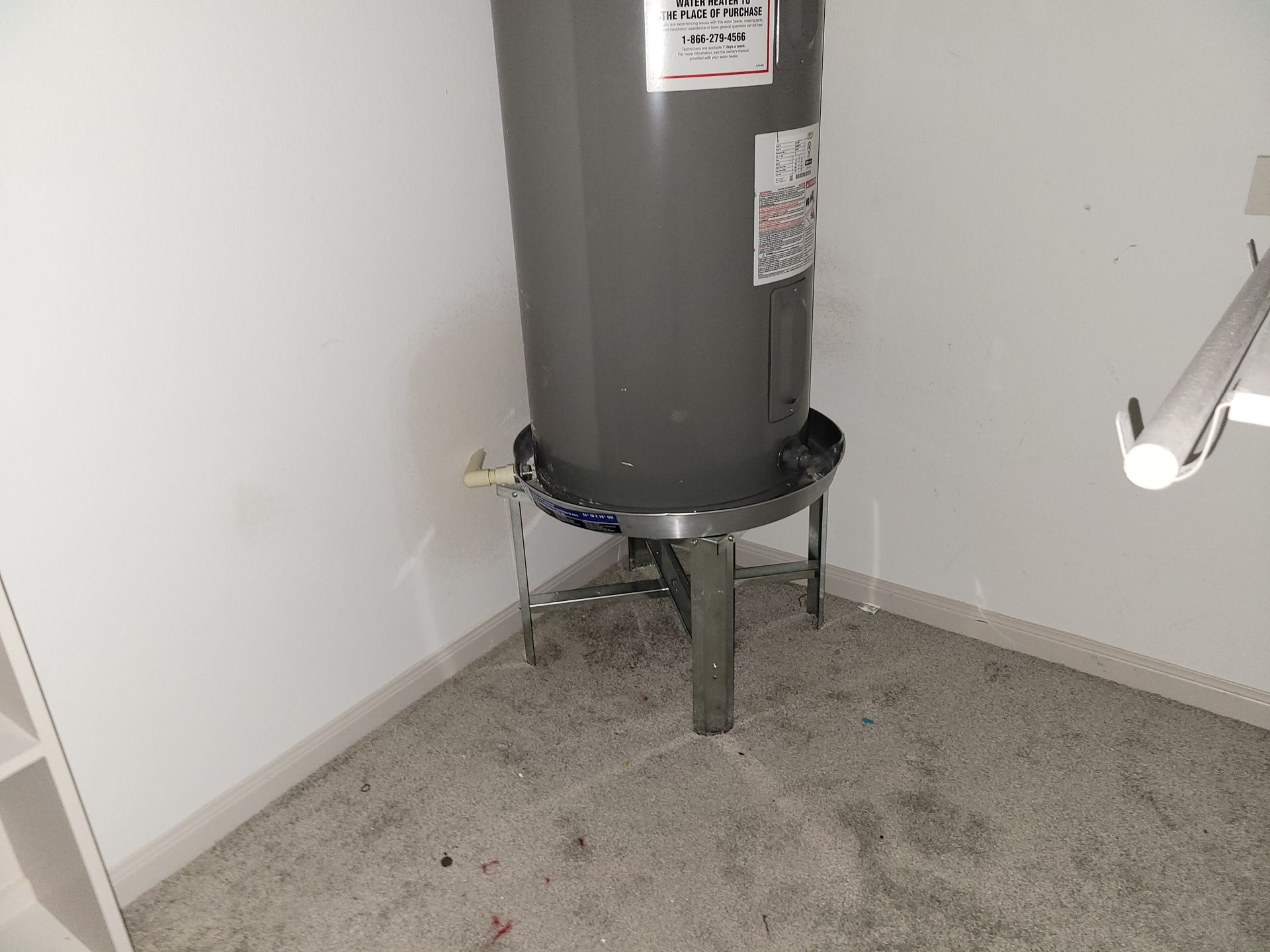 Water heater in a corner, gray tank on a metal stand with a drain pipe.
