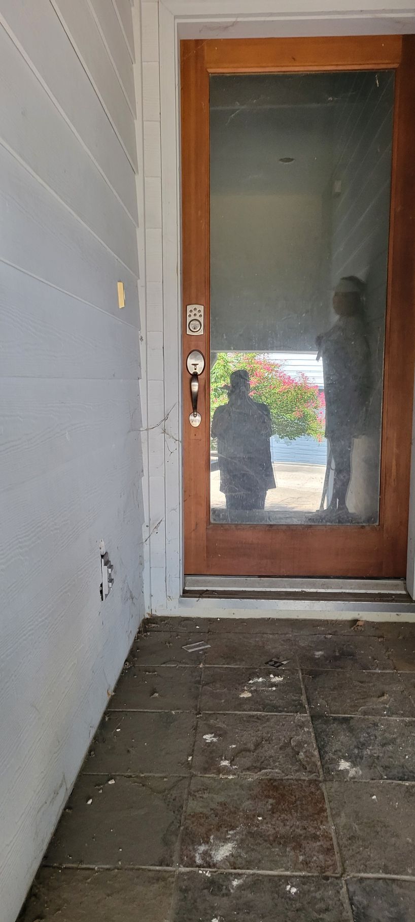 A wooden door with a large glass pane reflecting two people and a street view; the entrance has tiled flooring.