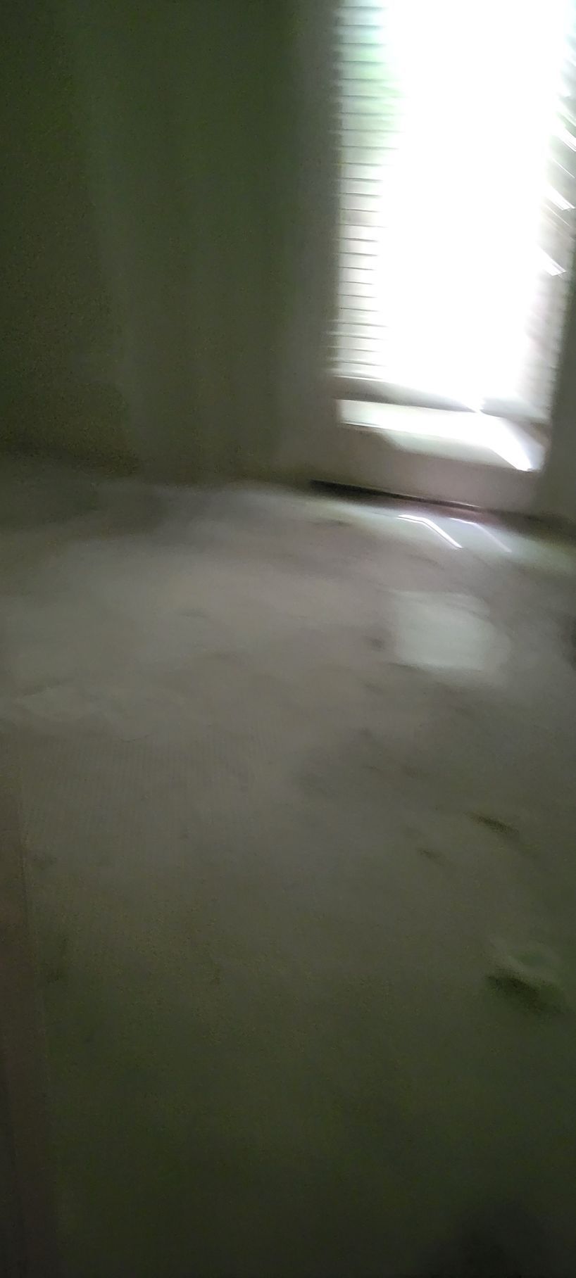 Empty, dimly lit room with a window covered by blinds; pale walls and floor; shadows and light.