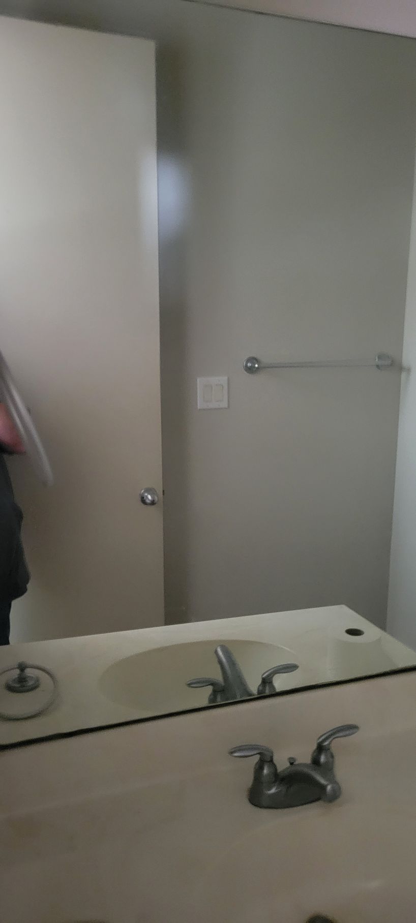 Bathroom mirror reflecting a sink, faucet, towel bar, and a wall with a light switch.