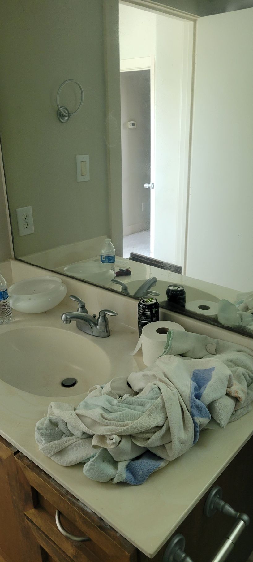 Bathroom sink with mirror, towels, and various items on the countertop. Light walls and door in the background.