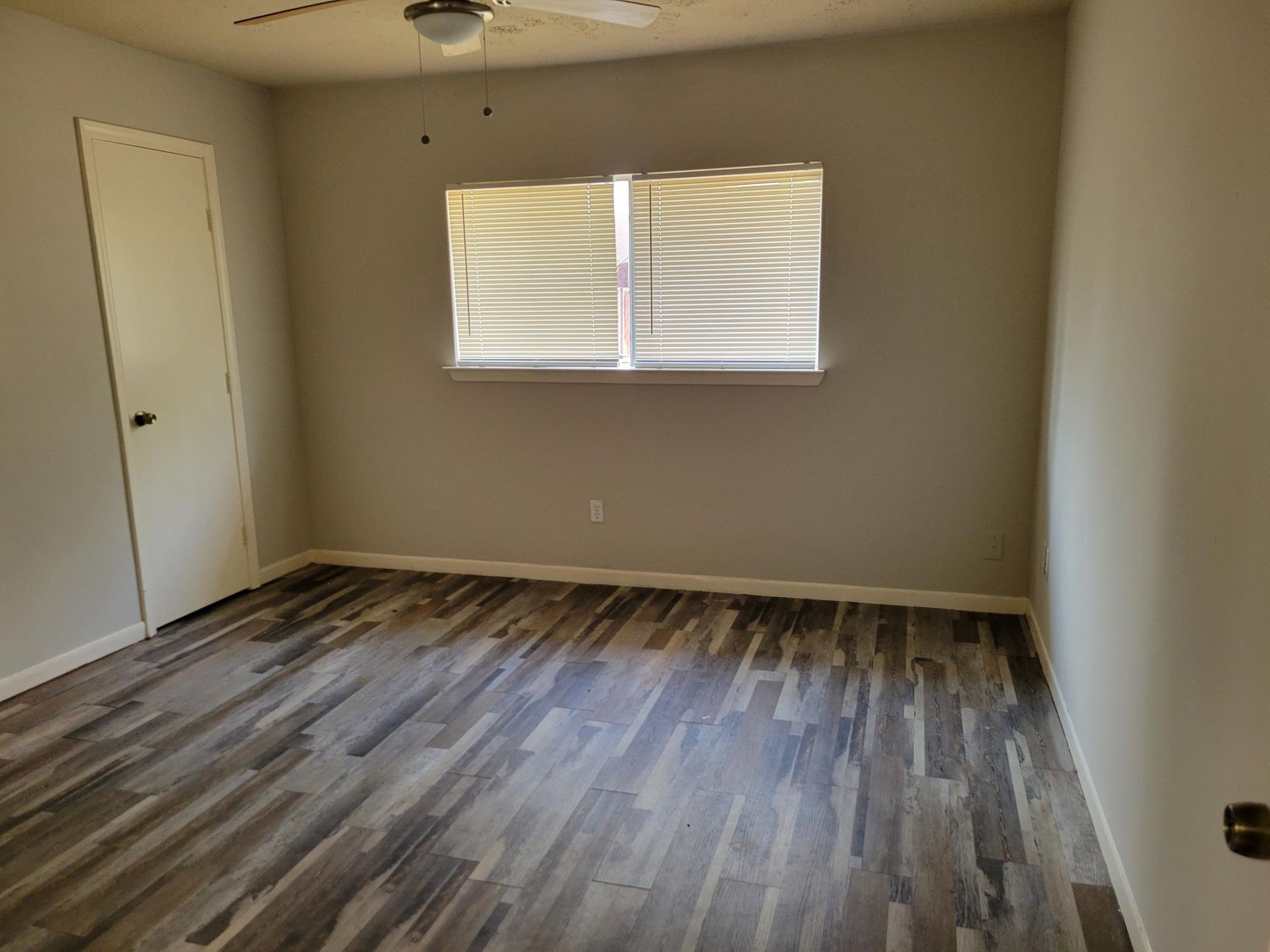 Empty bedroom with gray walls, wood-look flooring, window with blinds, and a closed door.