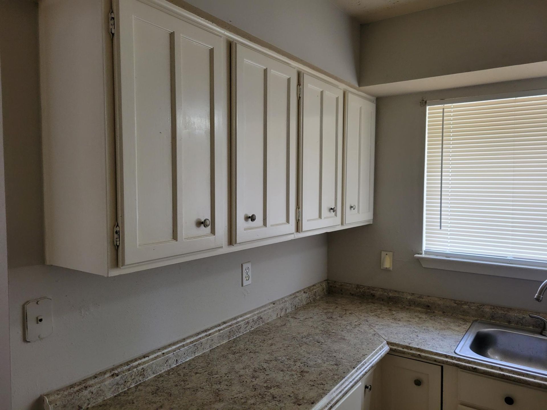 White kitchen cabinets over a speckled countertop and a window with blinds.