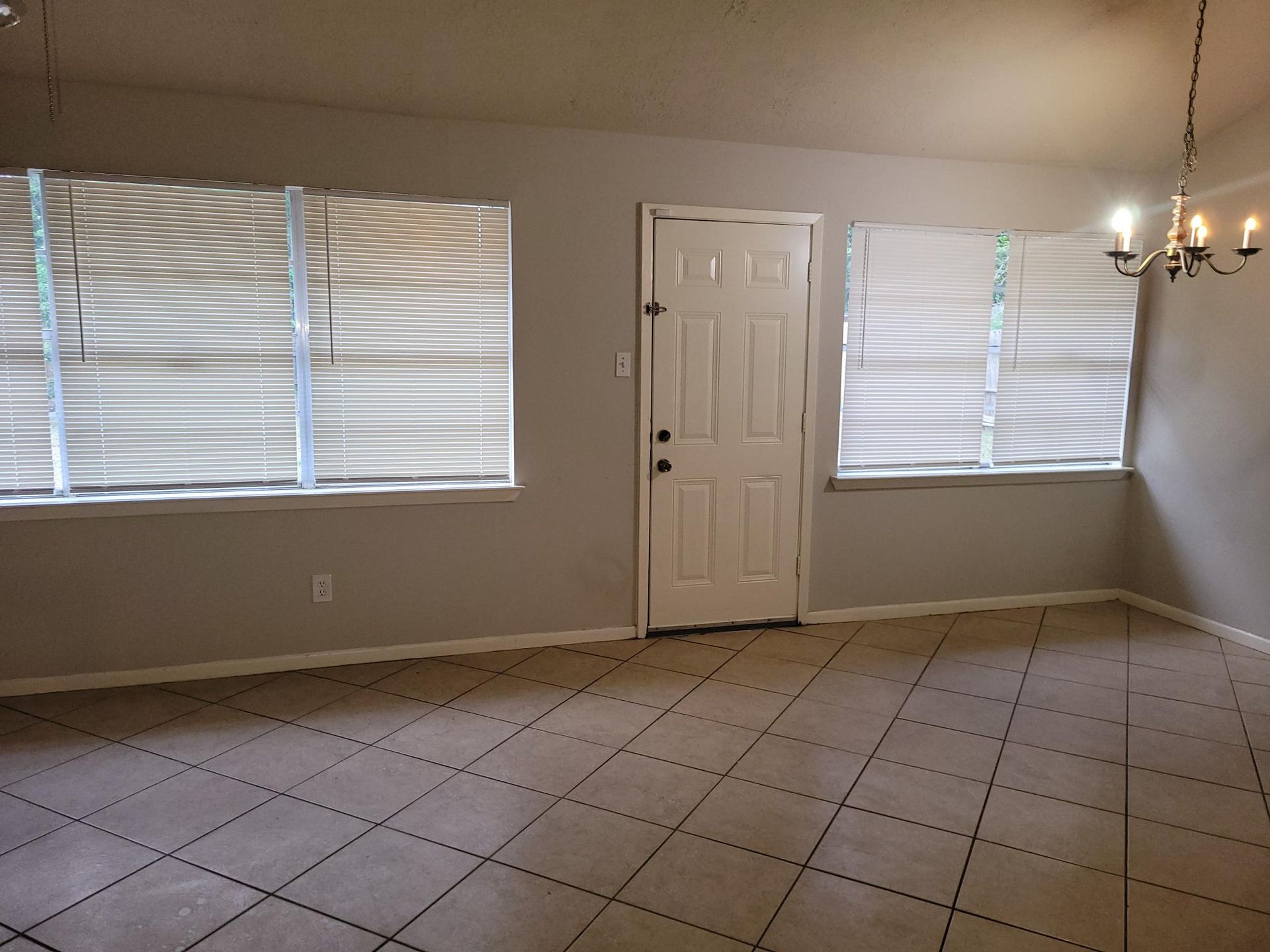 Empty room with gray walls, tiled floor, white blinds, and a chandelier.