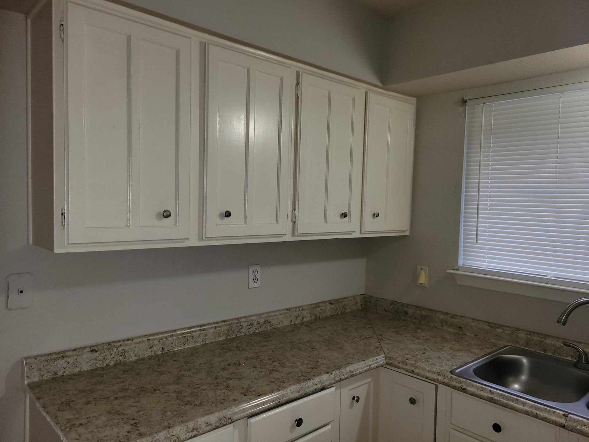 White kitchen cabinets above a speckled countertop, near a window with blinds and a sink.