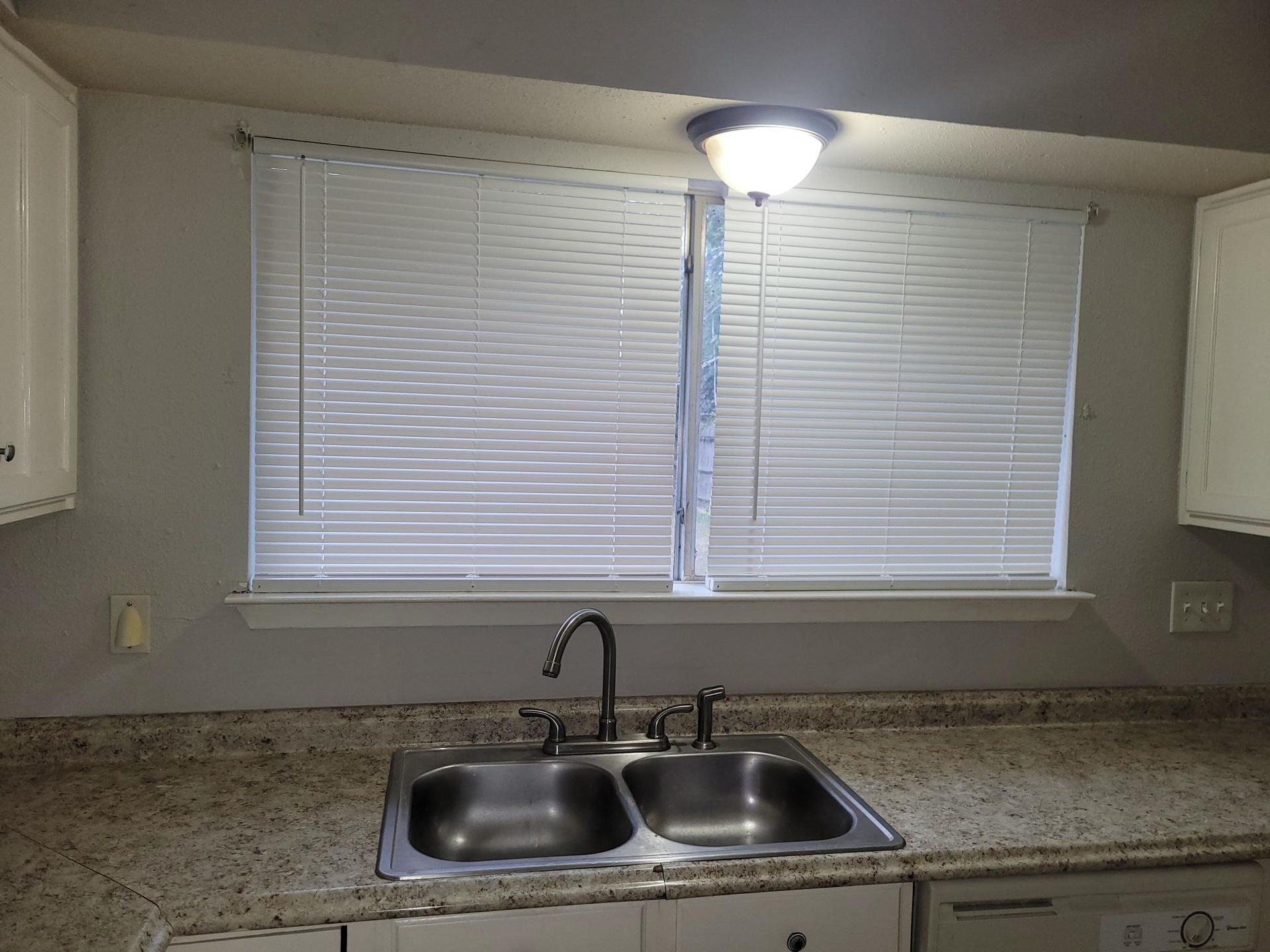 Kitchen sink under two windows with closed blinds. Overhead light. White cabinets and countertop.