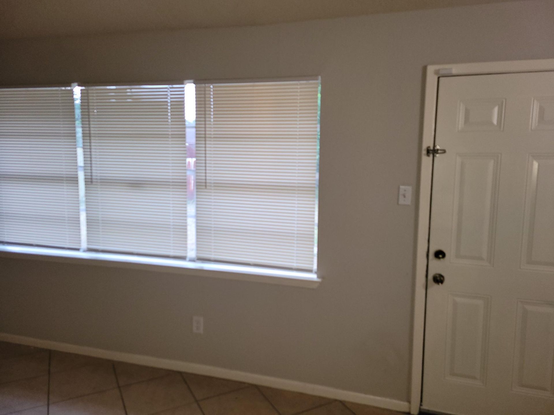 Room with a window and closed blinds, and a white door to the right. Walls are painted gray.
