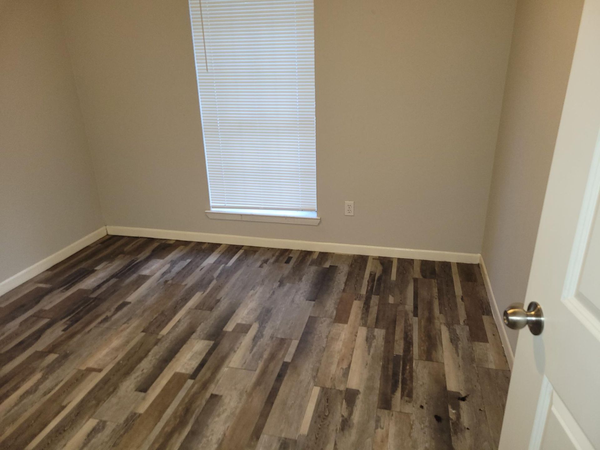 Empty room with wood-look flooring, beige walls, and a window with blinds.
