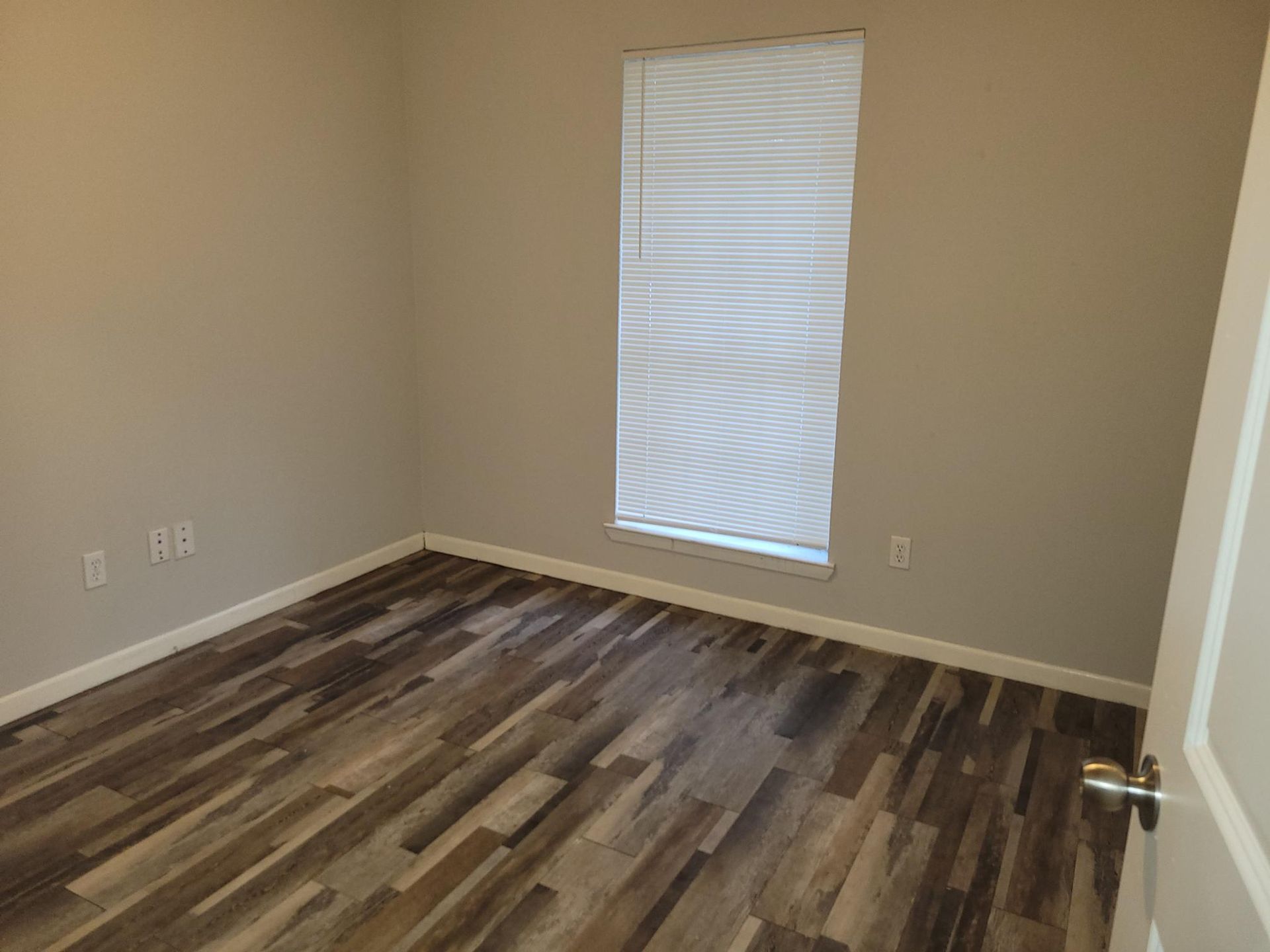 Empty room with wood-look flooring, light-colored walls, window with blinds, and a closed door.