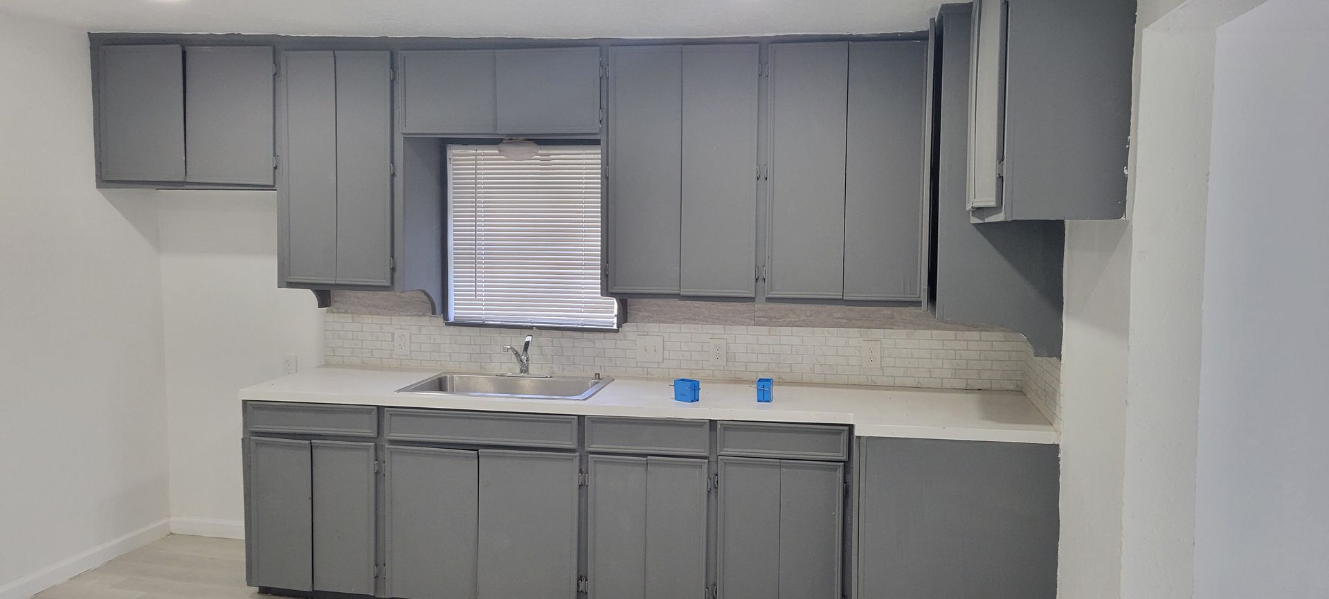 Gray kitchen cabinets, white countertops, a window, and a stainless steel sink.