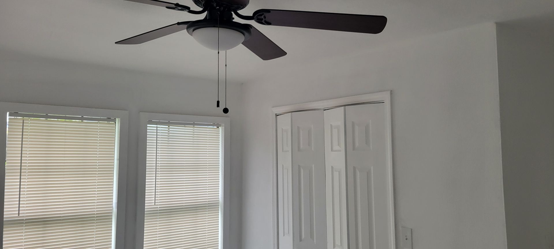 A ceiling fan, white closet doors, and a window with blinds. The walls are white.