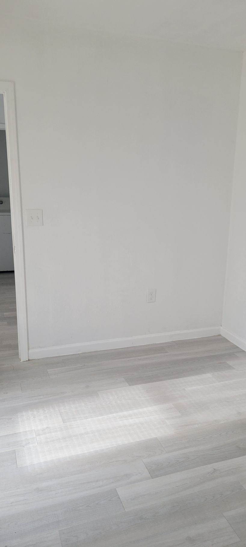 A room with light gray wooden floors and white walls; a doorway on the left.
