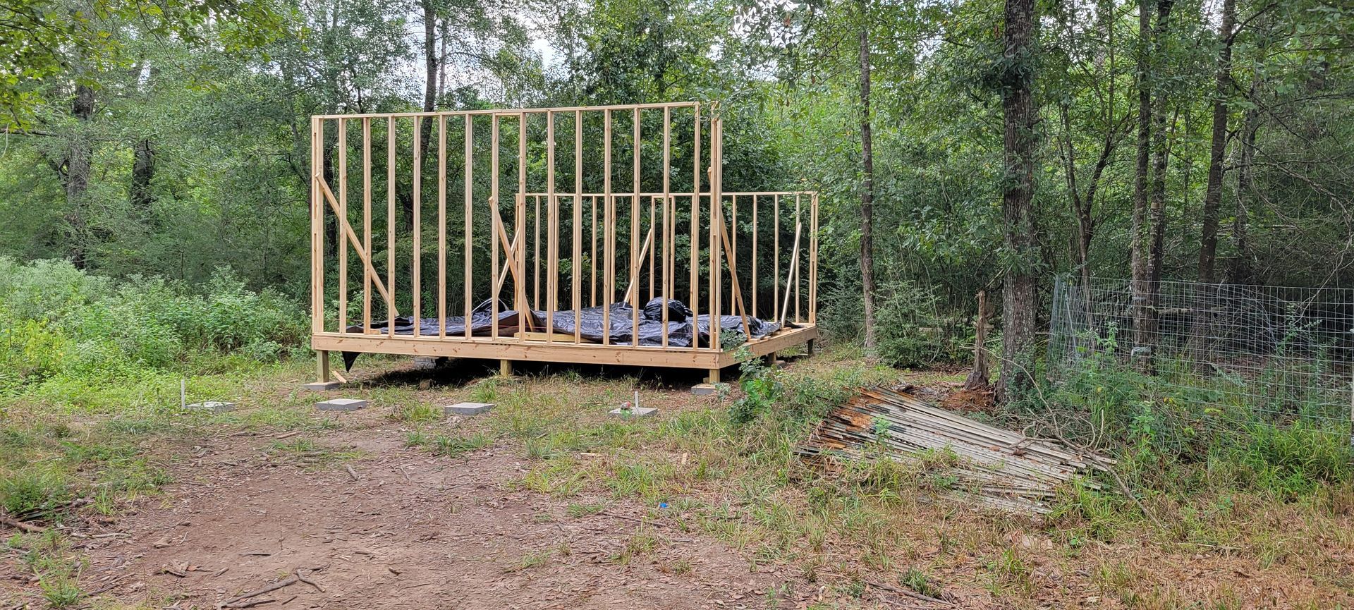 Wooden frame of a small building under construction, in a grassy clearing surrounded by trees.