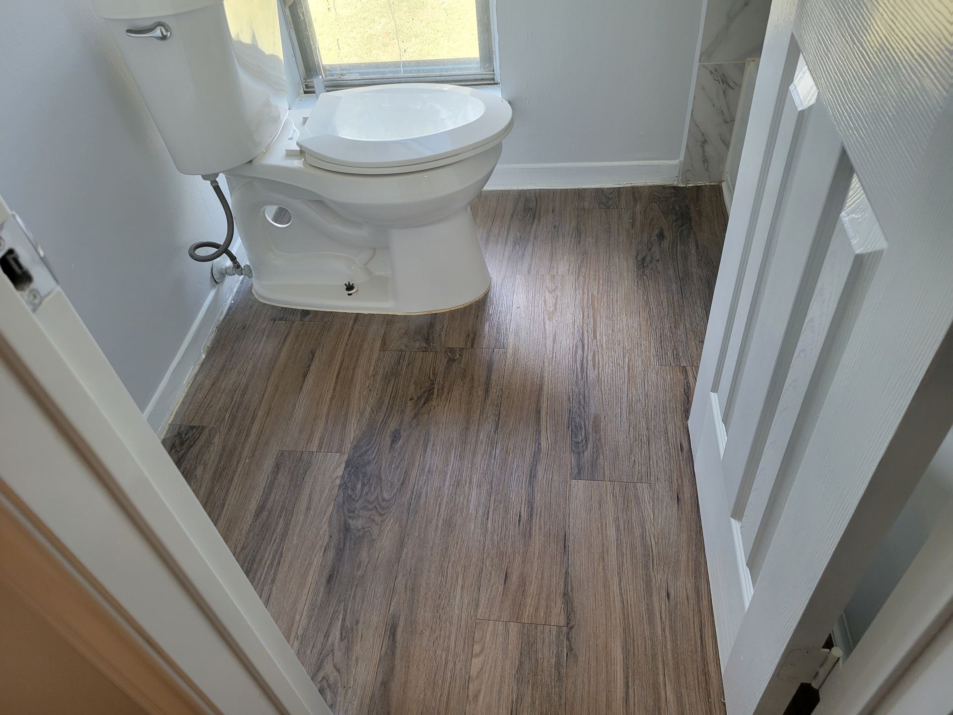 Toilet in a bathroom with wood-look flooring and a small window. White walls and door frame.
