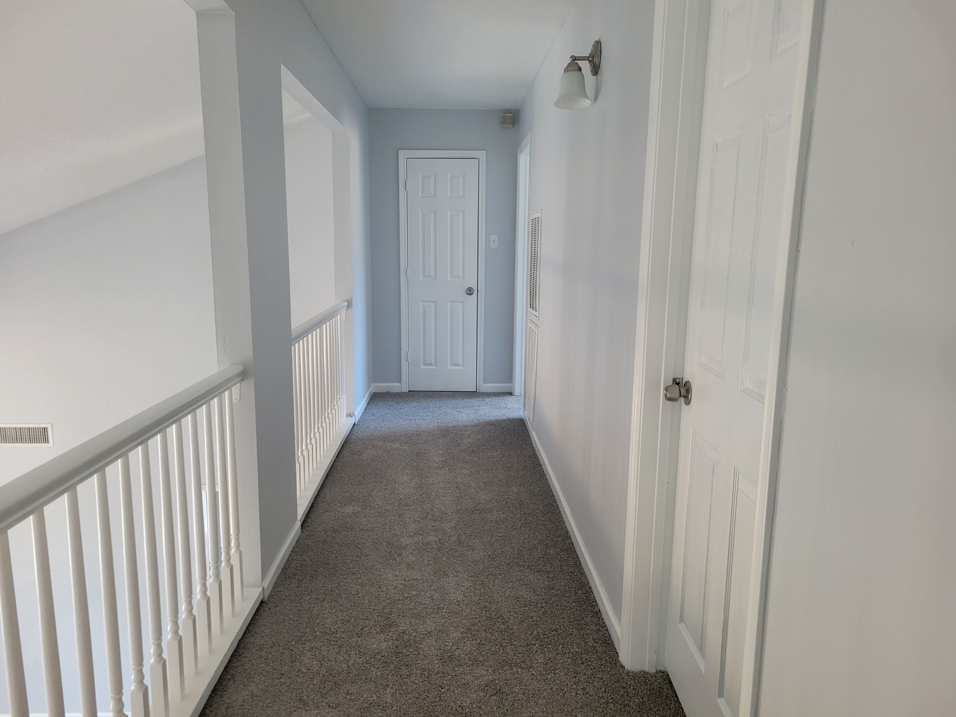 Hallway with gray carpet, white walls, and a door at the end. A railing is on the left.