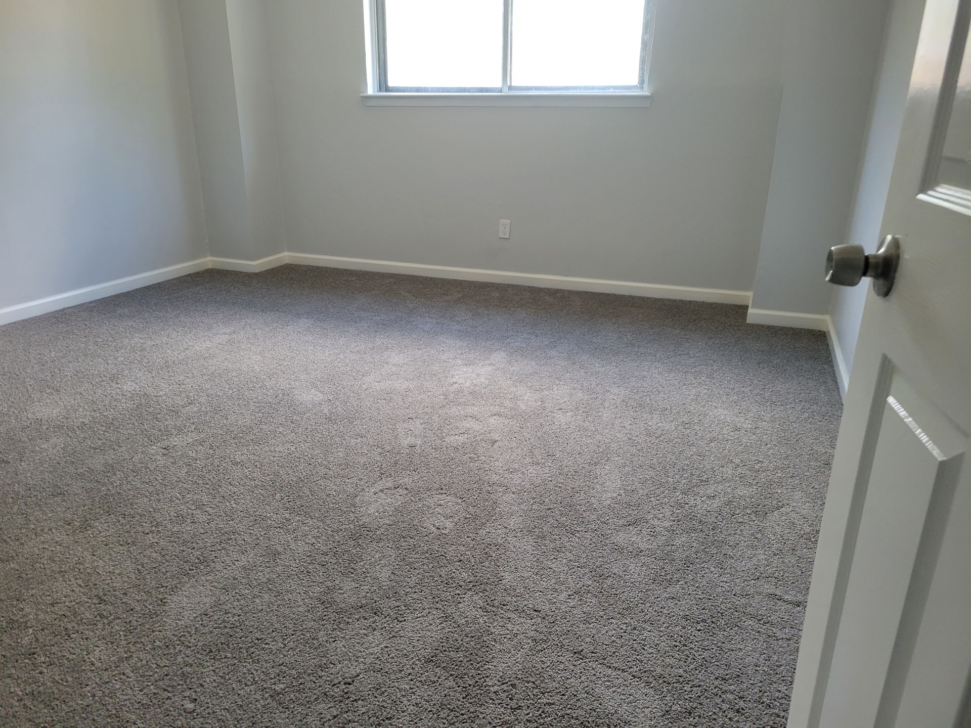 Empty room with gray walls and carpet, a window, and a partially open white door with a doorknob.