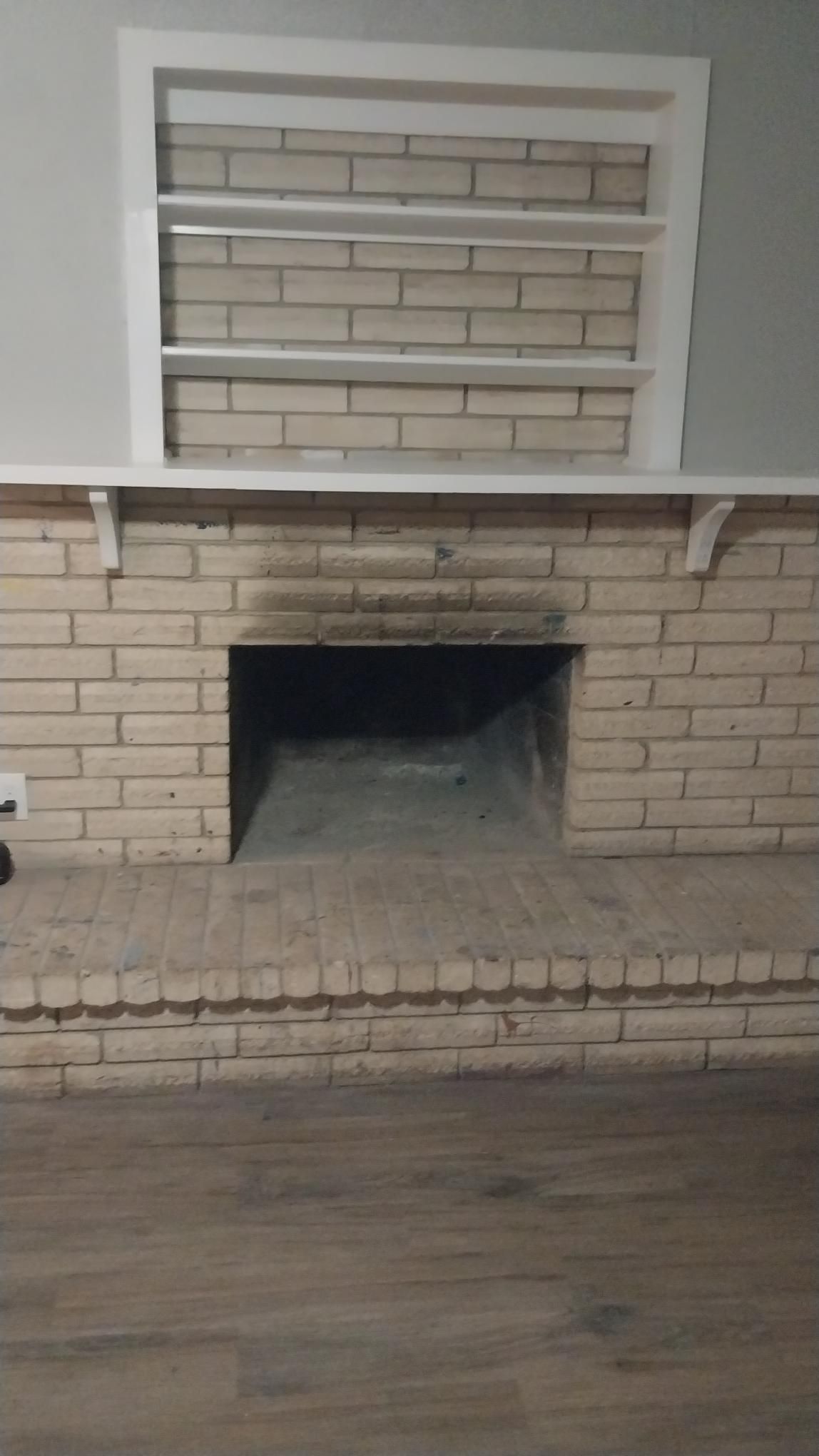 Brick fireplace with white mantel and shelves, dark interior, light brick exterior, and wood floor.