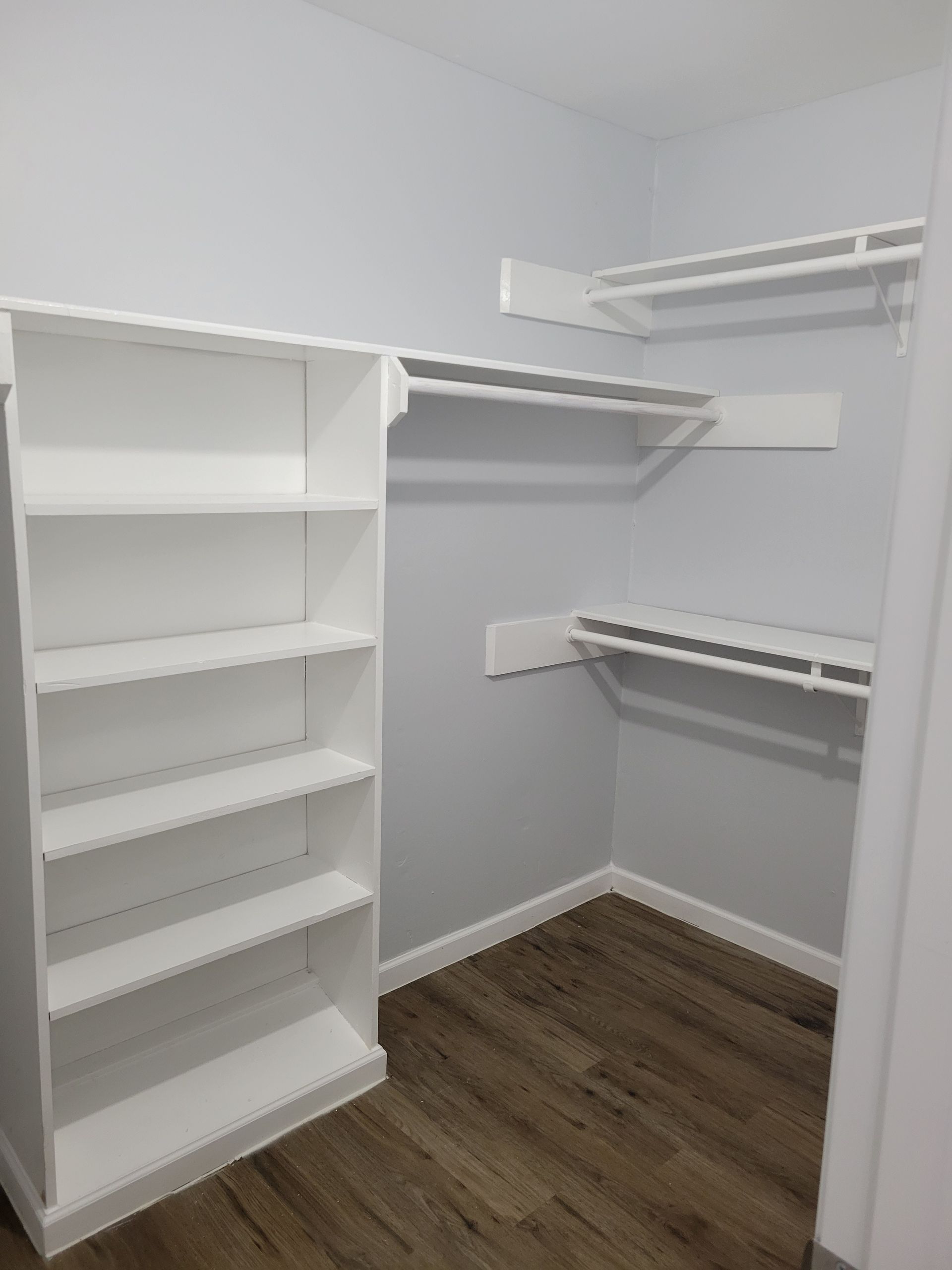 Empty walk-in closet with white shelving and rods, gray walls, and wood-look flooring.