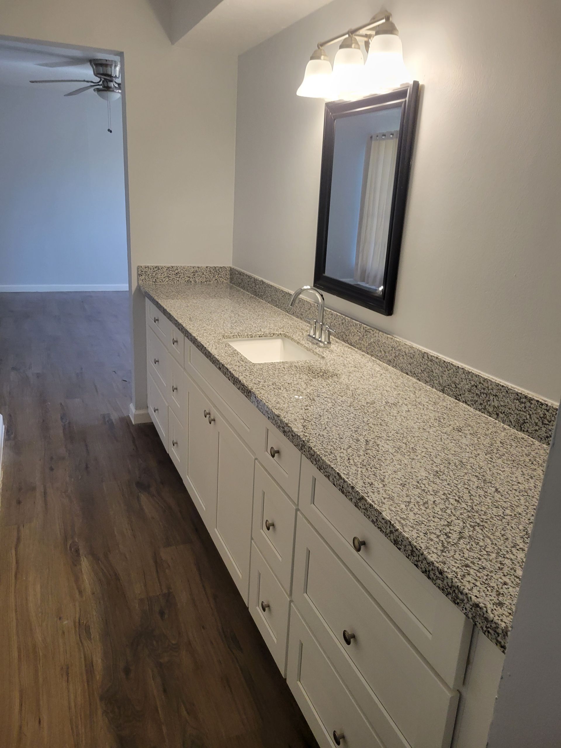 Bathroom with long white vanity, granite countertop, mirror, and hardwood floor.