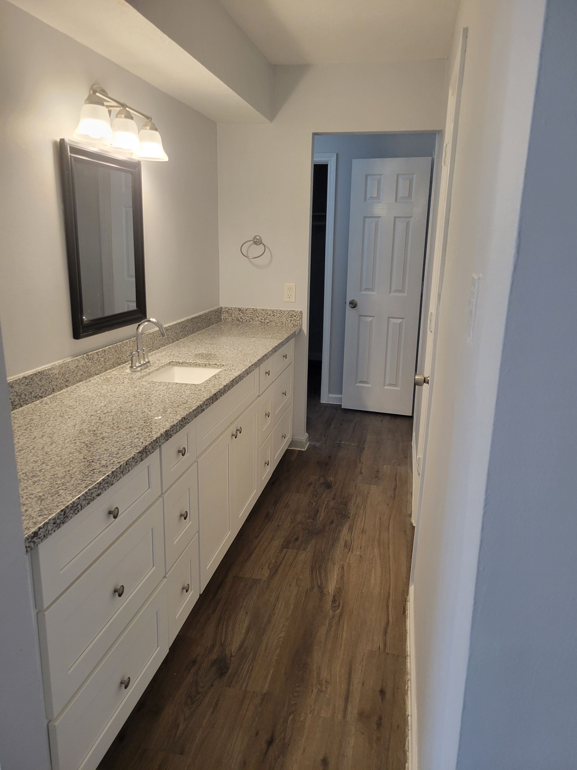 Bathroom with long white vanity, granite countertop, mirror, and a door at the end of the hallway.
