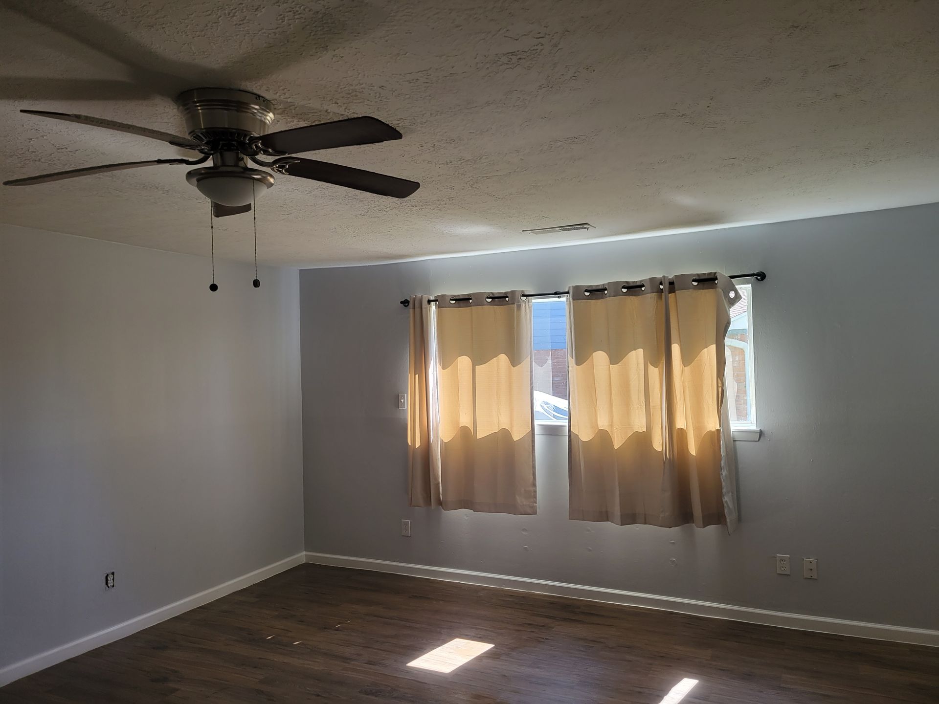 Empty room with a ceiling fan, window with curtains, and wood-look flooring. Sunlight streams in.
