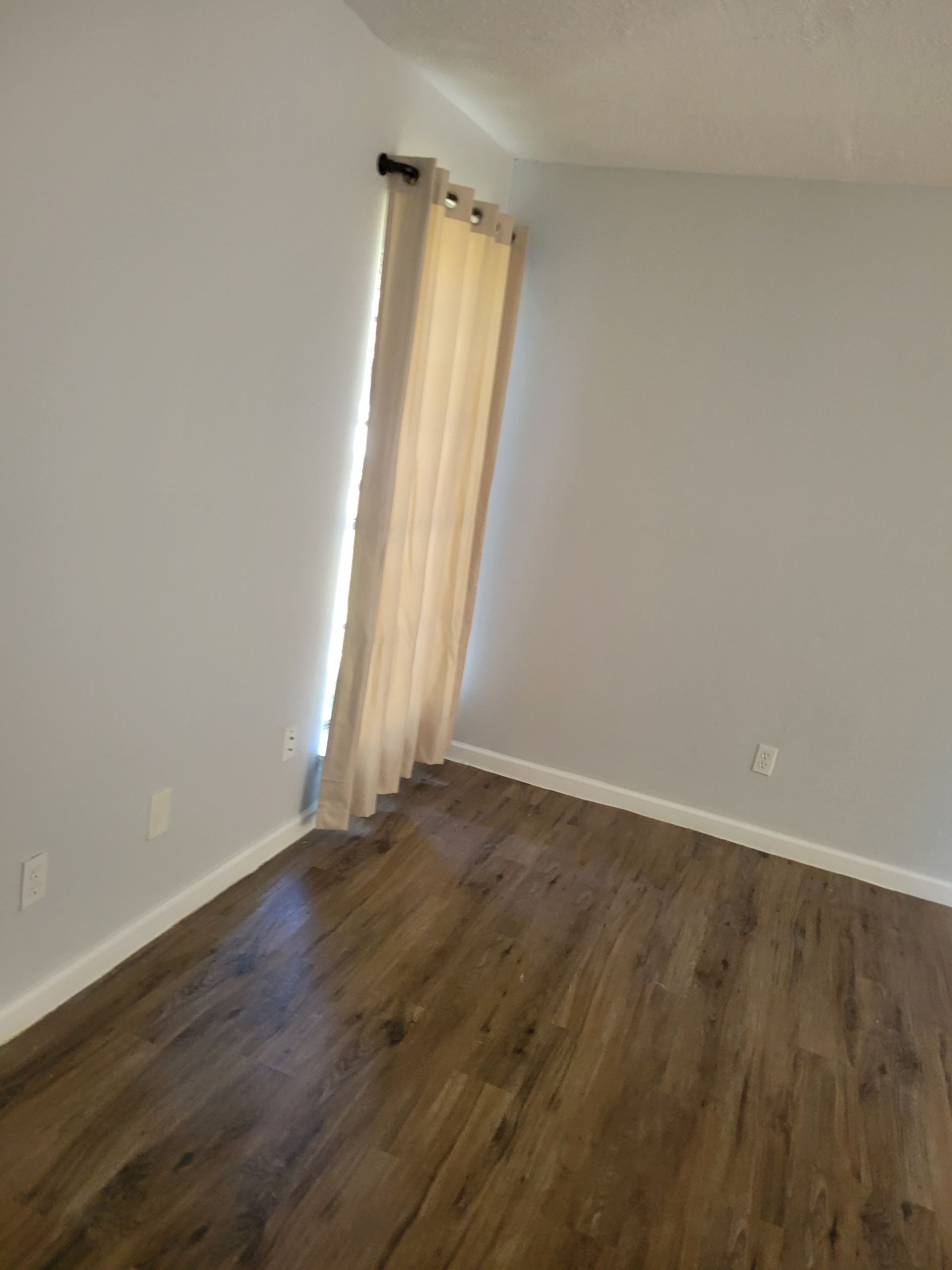 Empty room with light gray walls, dark wood-look floor, and a beige curtain.