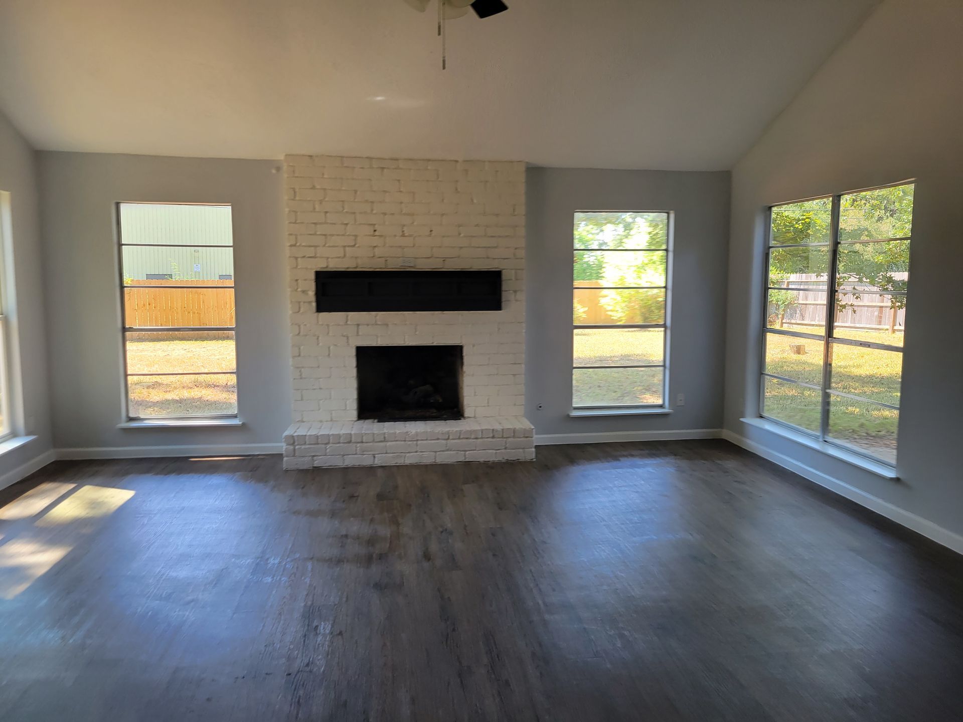 Empty living room with painted brick fireplace, dark wood-look floor, and windows.