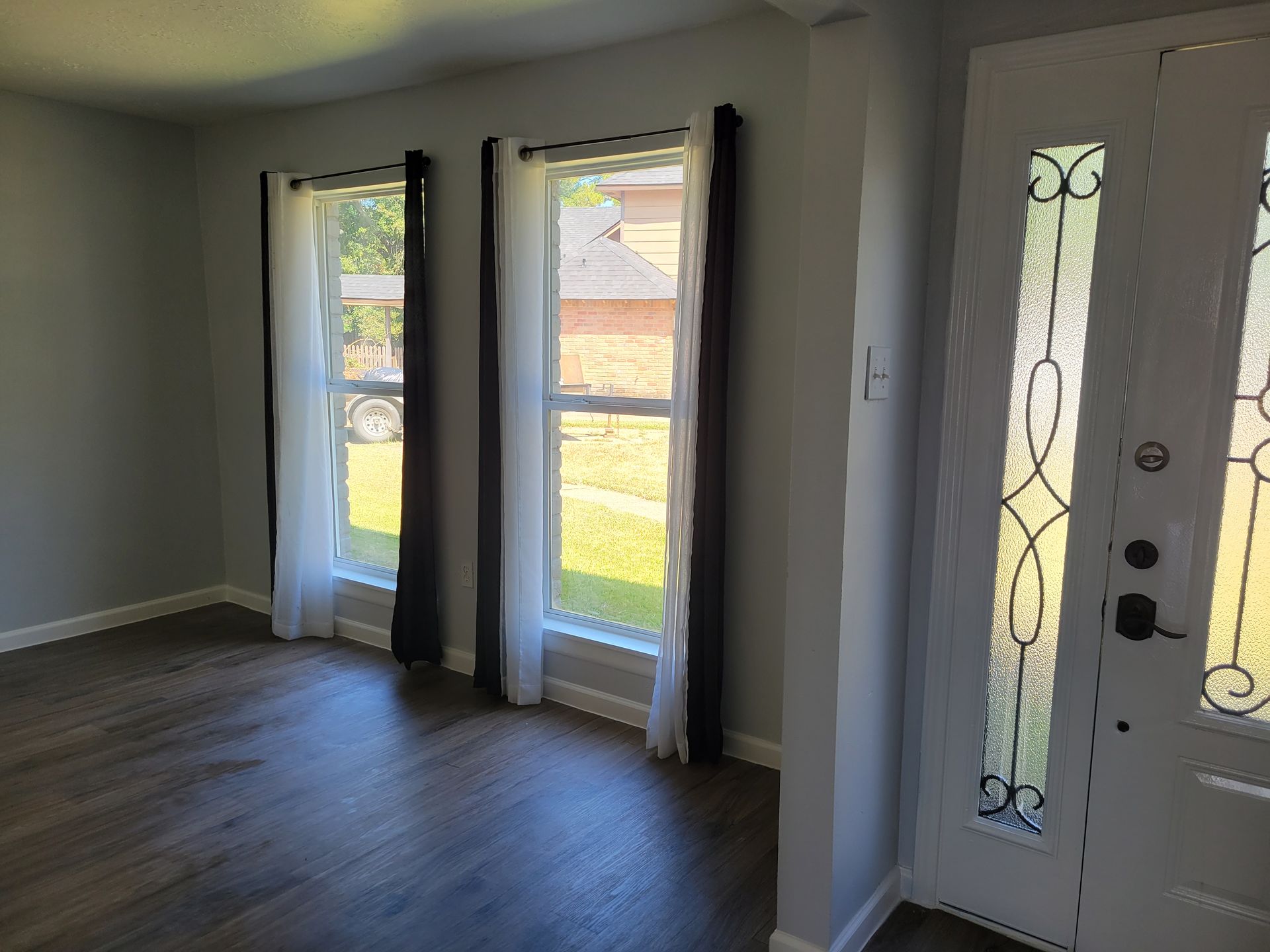 Interior room with two windows and a front door. Gray walls, dark curtains, and wood-look flooring.