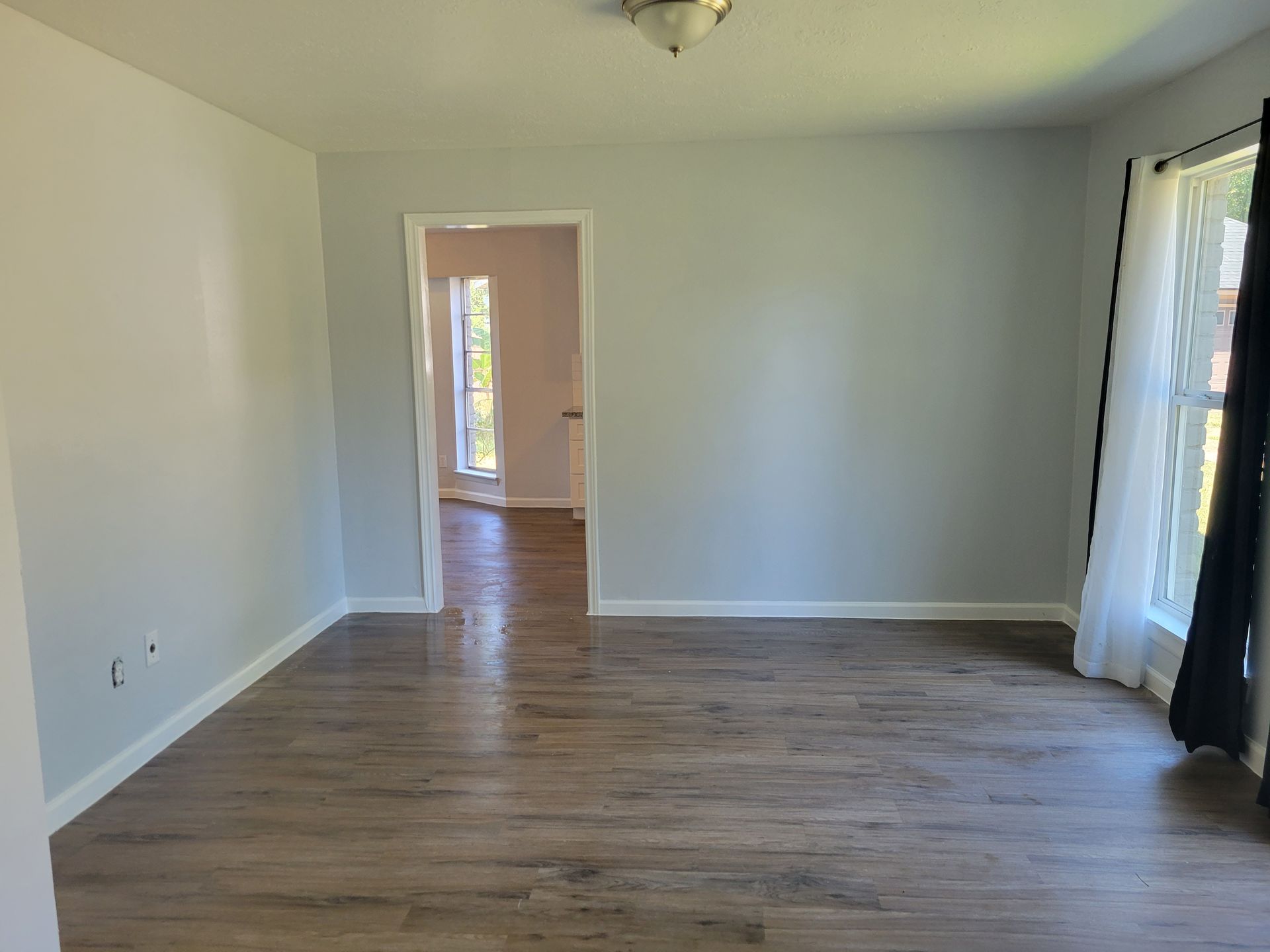 Empty room with gray walls, wood-look flooring, and a doorway to another room. A window with curtains is on the right.