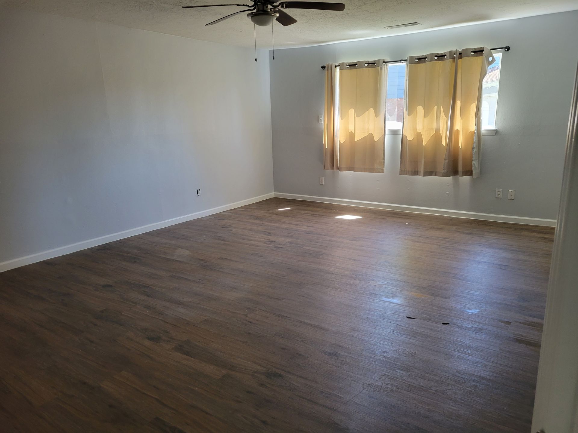 Empty room with wood-look flooring, light gray walls, and a window with beige curtains. A ceiling fan is visible.