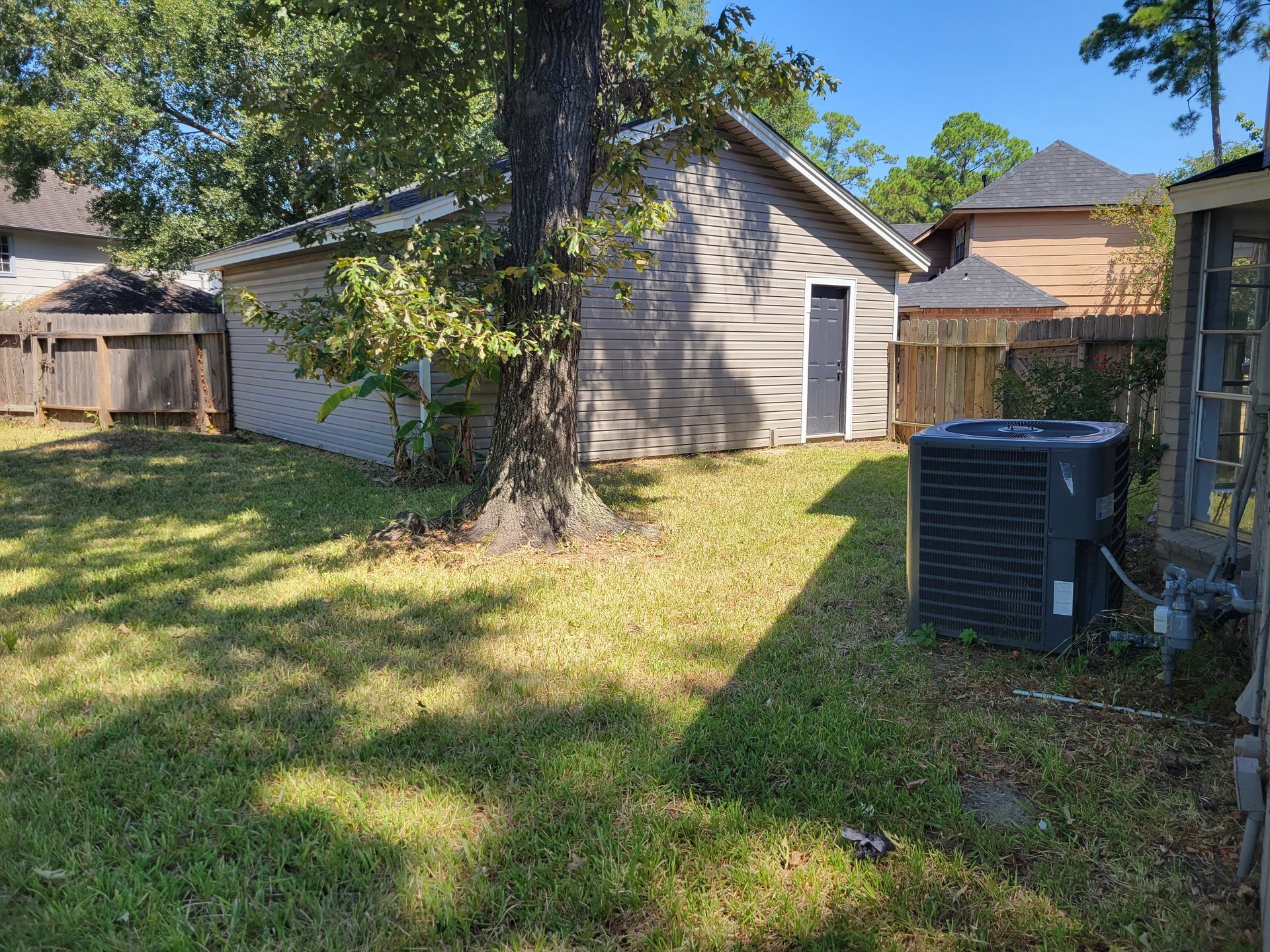Backyard with a detached garage, tree, and air conditioning unit. Fenced on both sides. Overcast day.