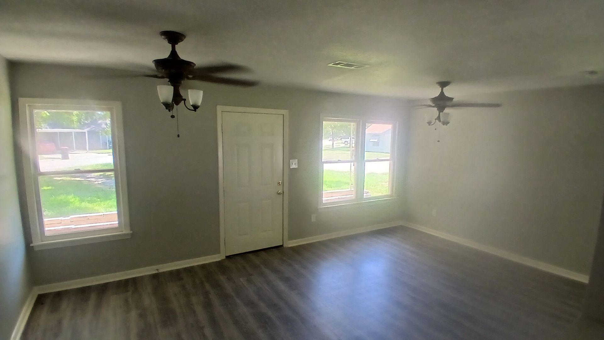 An empty living room with gray walls, wooden flooring, two ceiling fans, and windows.