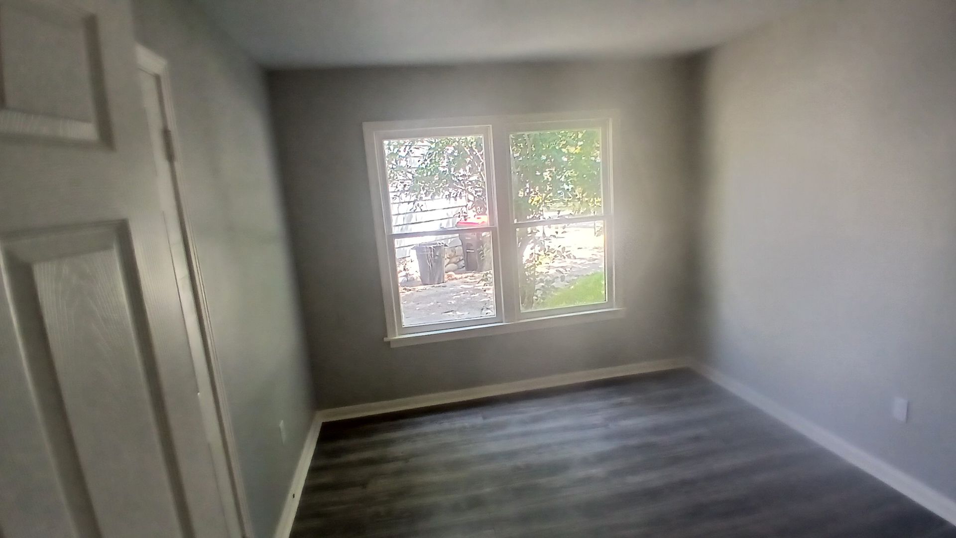Empty gray-walled room with a window and dark wood-look flooring; a white door is visible on the left.