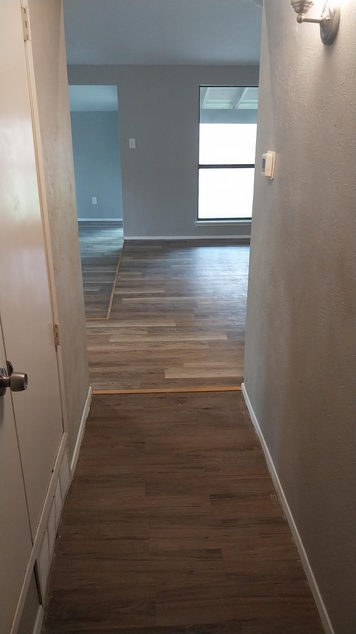 Narrow hallway leading to a room with a large window and wood flooring. Light fixtures on right wall.