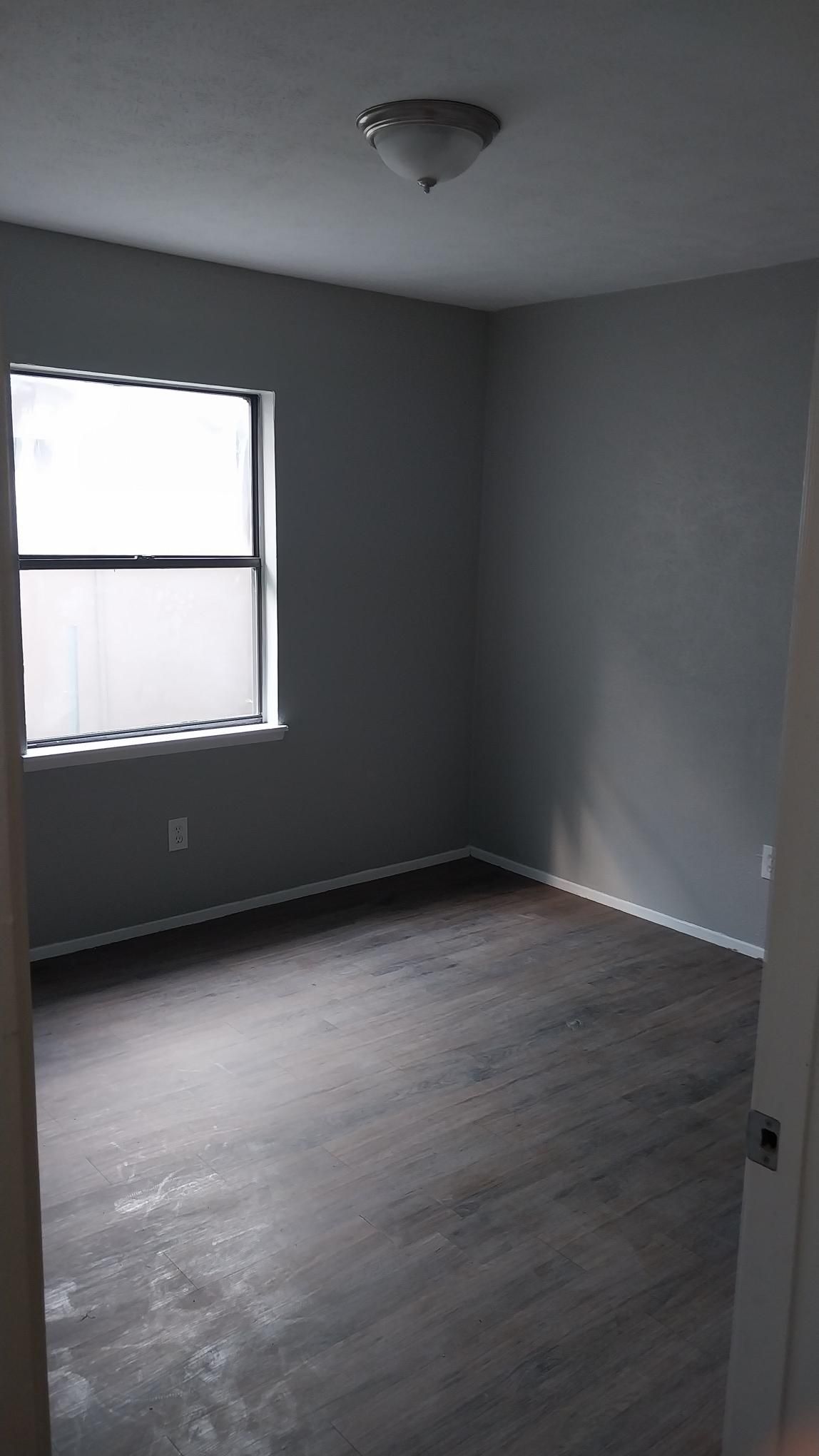 Empty room with gray walls, dark wood-look floor, and a window. A light fixture is on the ceiling.