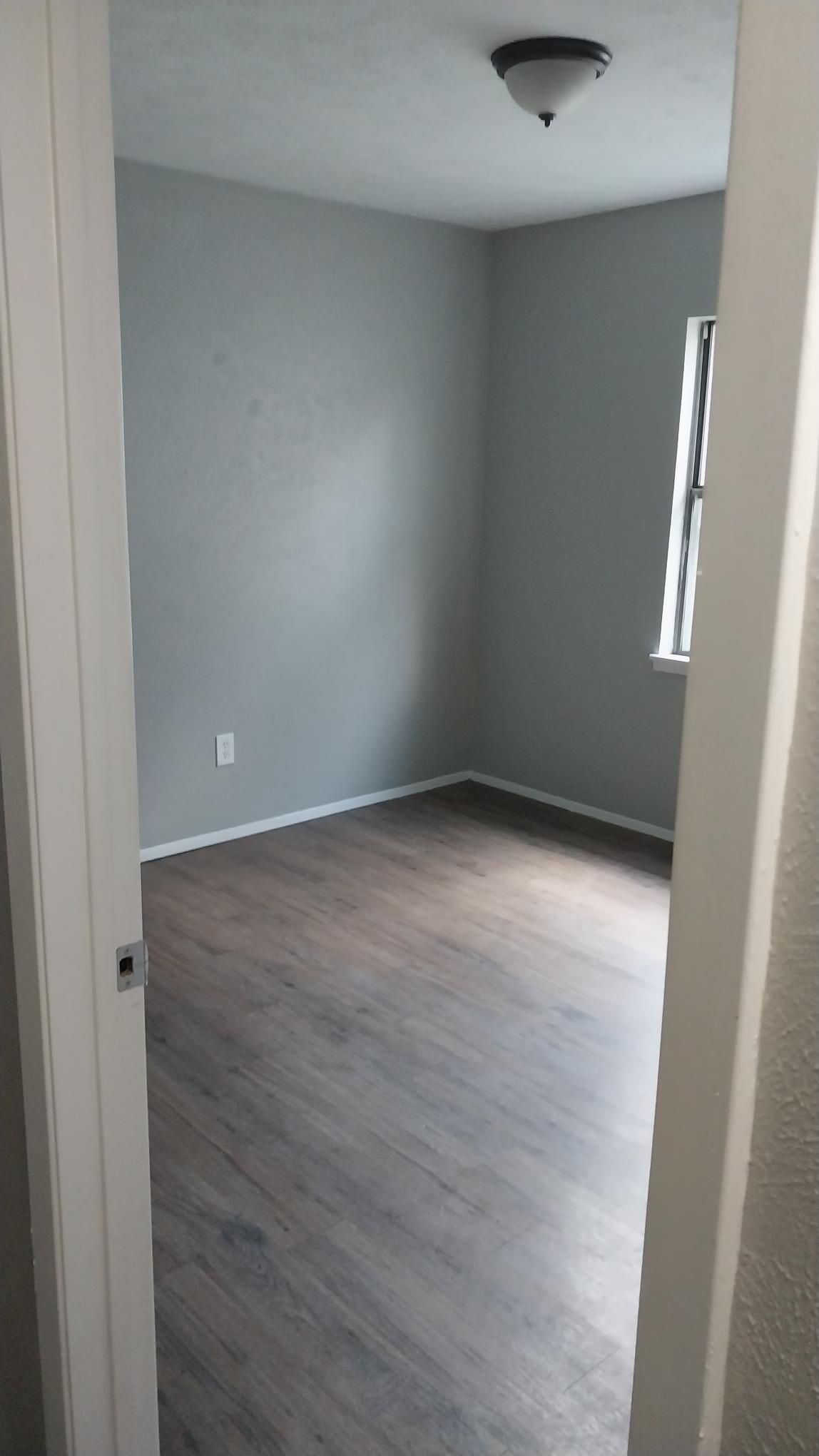 Empty bedroom with gray walls, dark wood-look floor, and a small window.