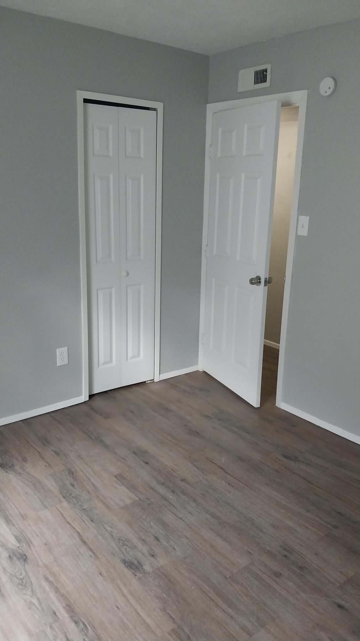 Empty bedroom with gray walls, wood-look flooring, and two white doors: one closed, one open to a hallway.