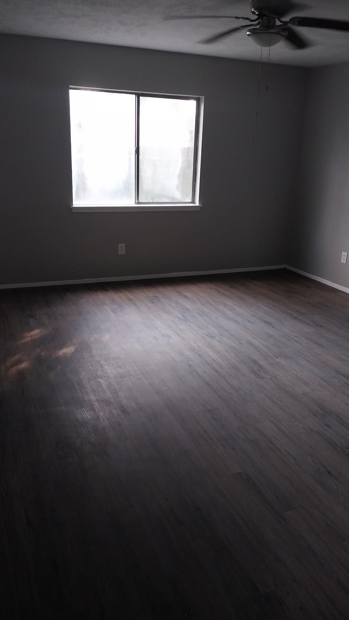 Empty room with dark wood flooring, gray walls, window, and ceiling fan.