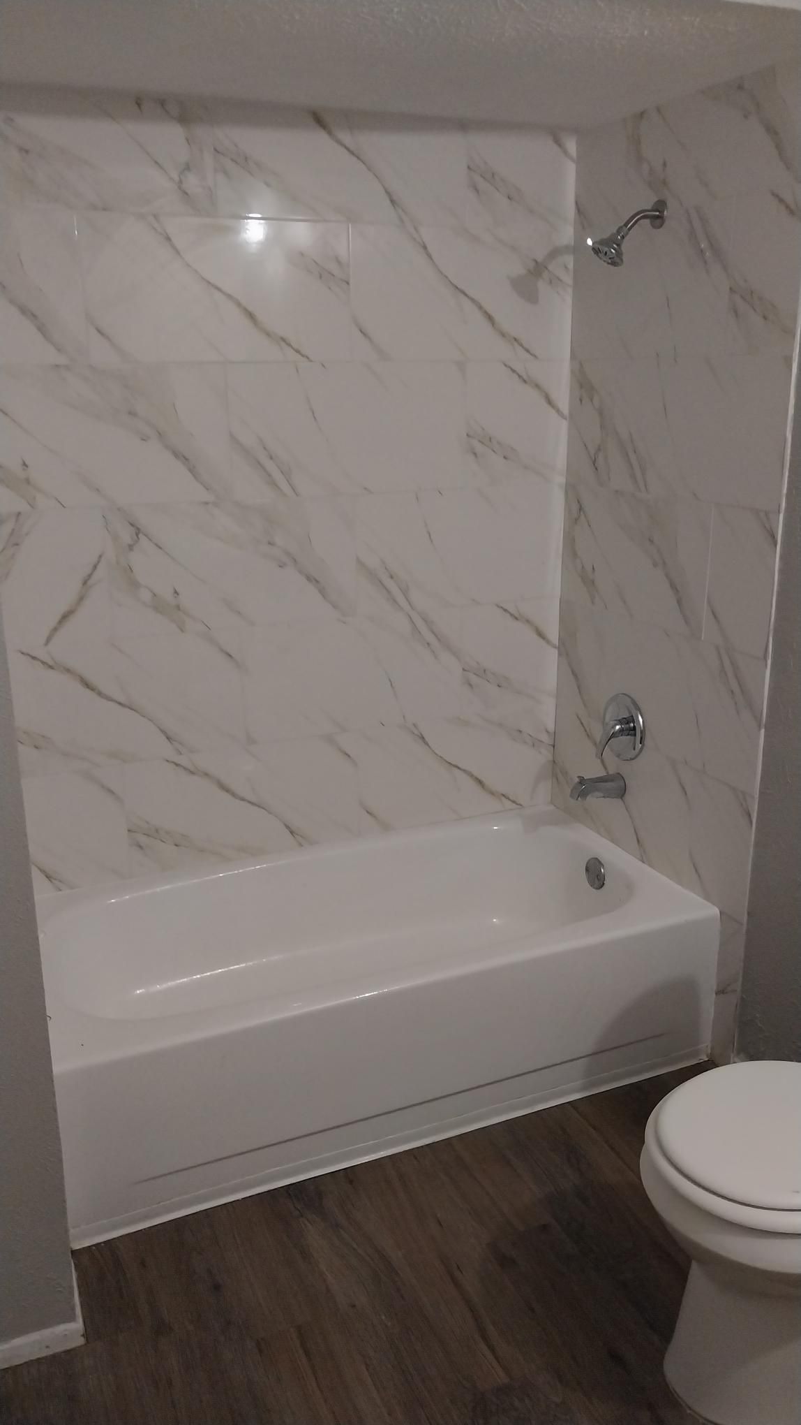 White bathtub and marble-look shower surround in a bathroom with dark wood-look flooring.