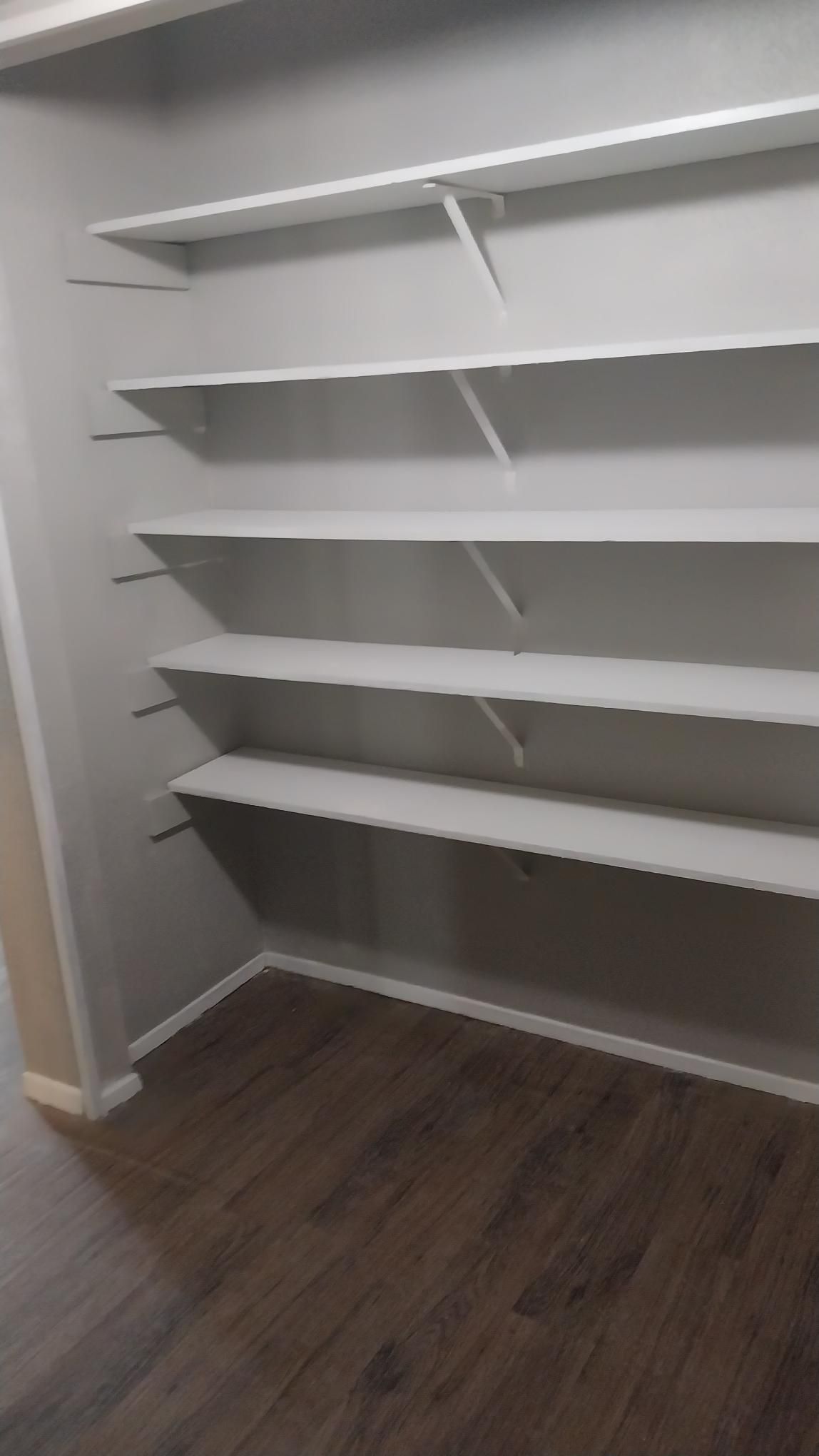 Empty closet with white shelves, gray walls, and wood floor.