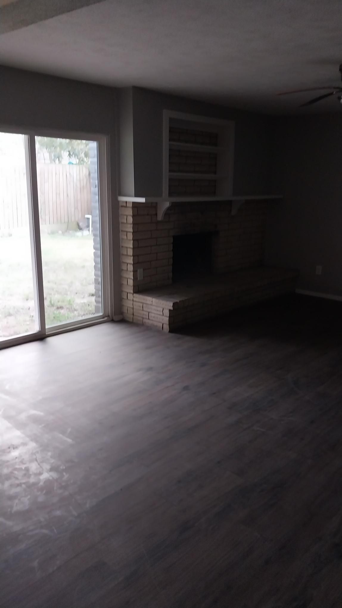 Living room with brick fireplace, sliding glass door to backyard, and dark wood-look flooring.