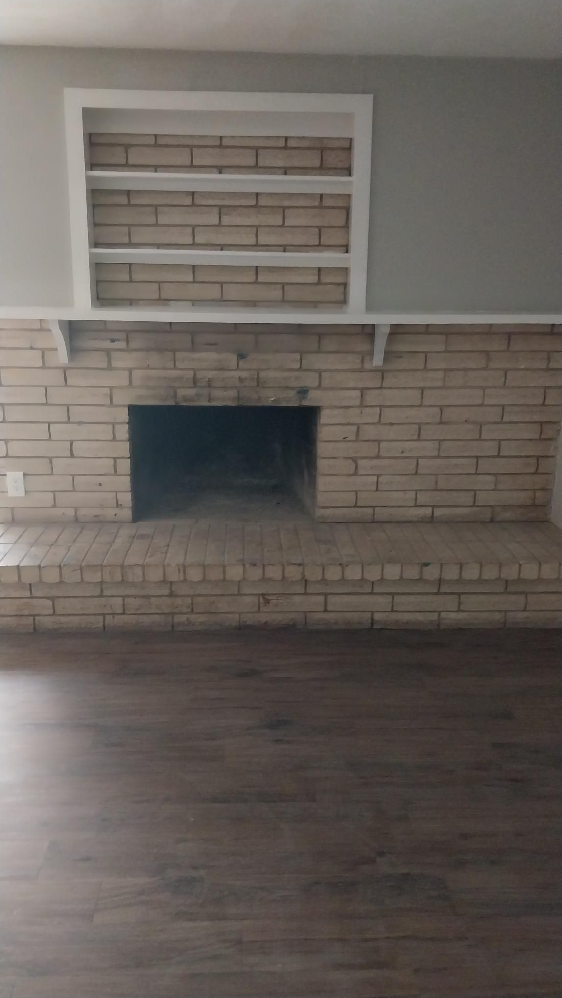 Brick fireplace with white built-in shelves and a raised hearth. Gray walls and wood-look flooring.