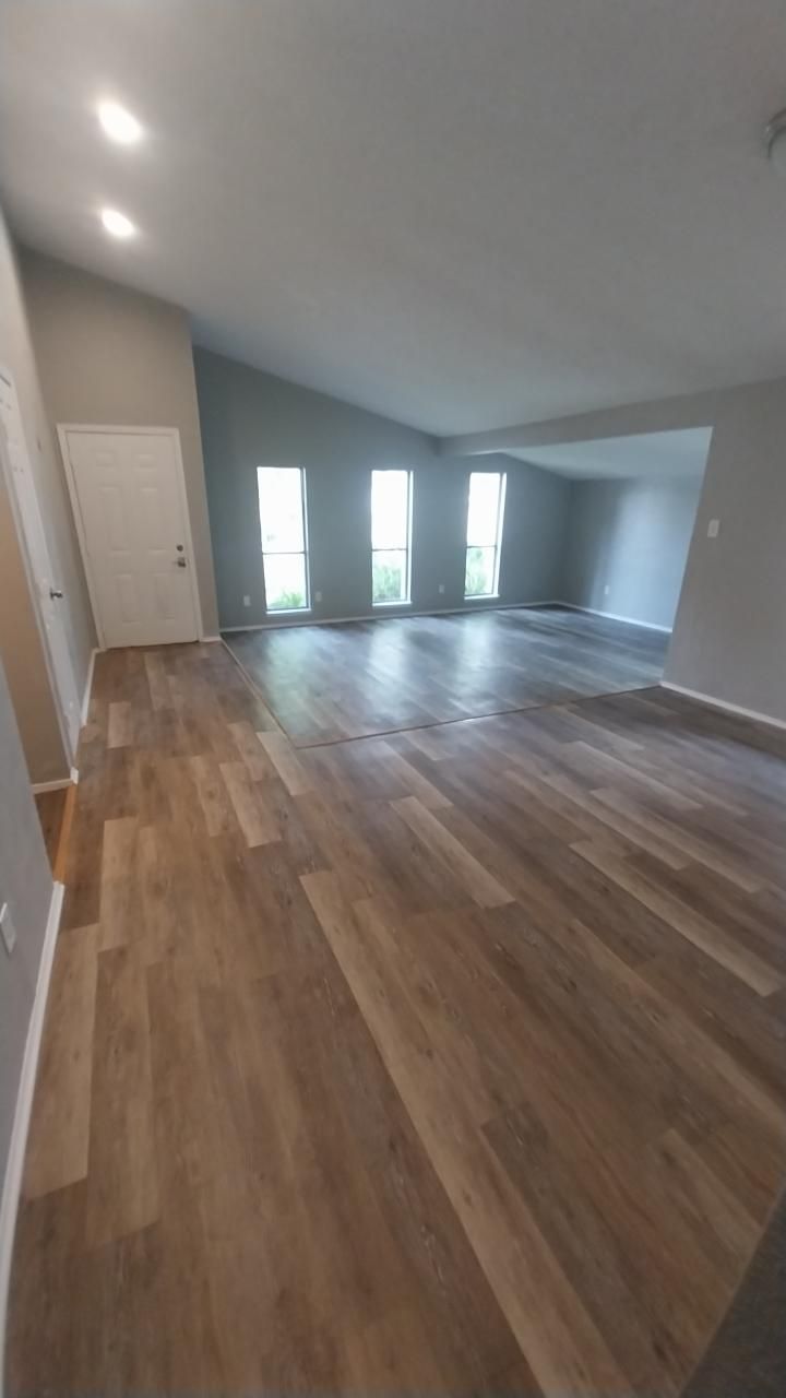 Empty living room with wood-look flooring, three tall windows, and a white door. Gray walls and recessed lights.