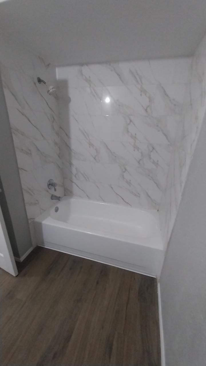 White marble-patterned shower walls over a white bathtub. Brown wood-look floor.