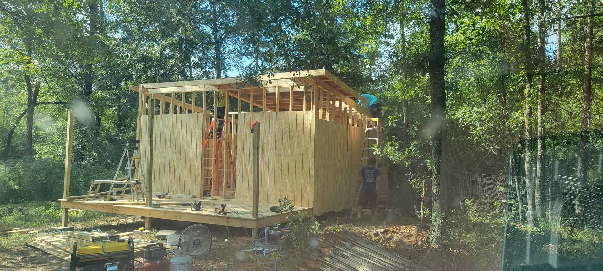 A wooden structure under construction in a wooded area, with trees in the background.