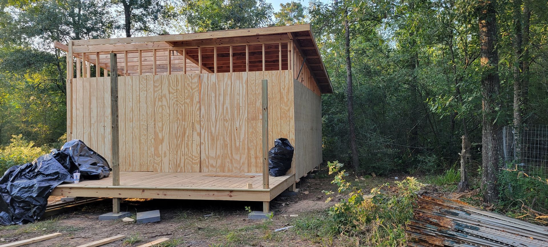 A wooden structure under construction in a wooded area, with OSB walls, a deck, and exposed framing.