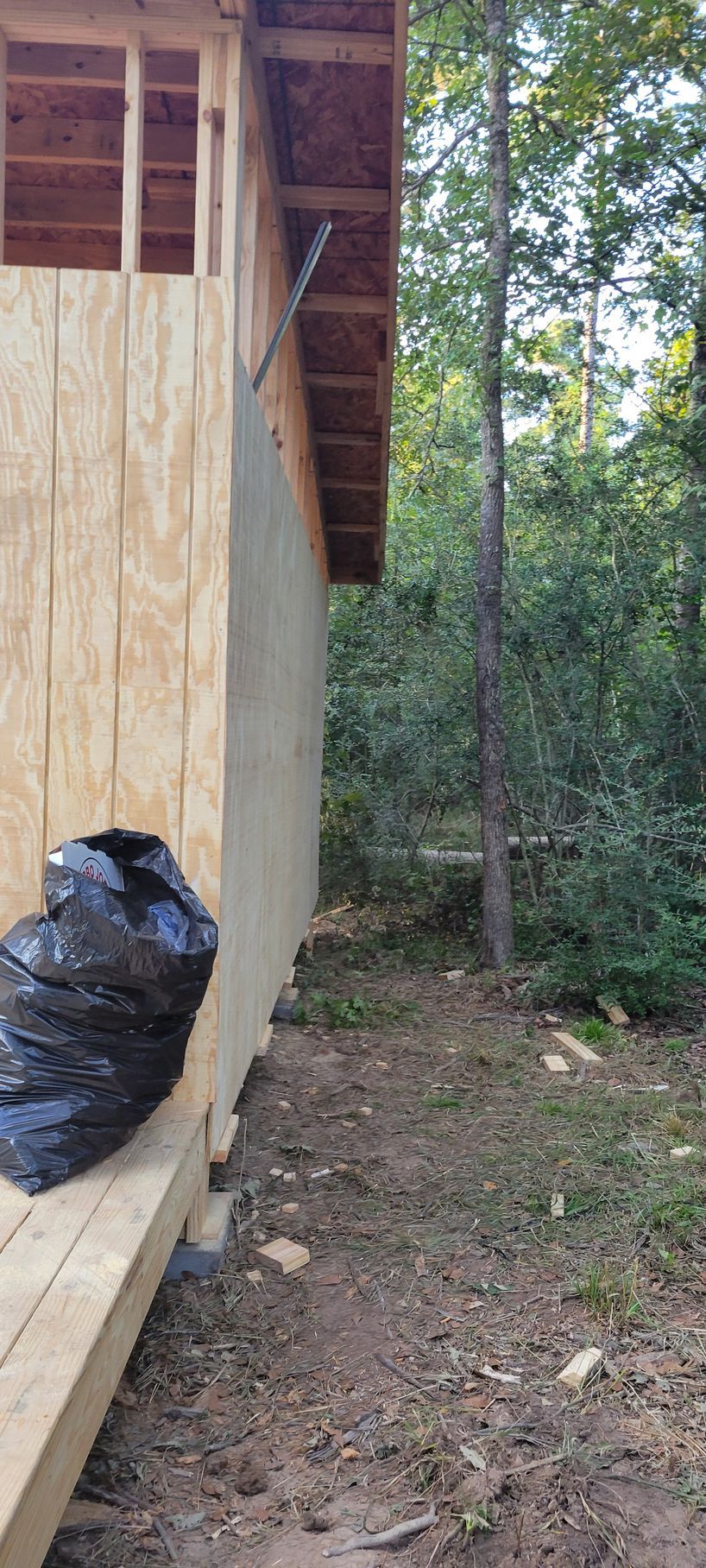 Side view of a partially constructed wooden building with a black trash bag on a wooden deck next to dirt and trees.