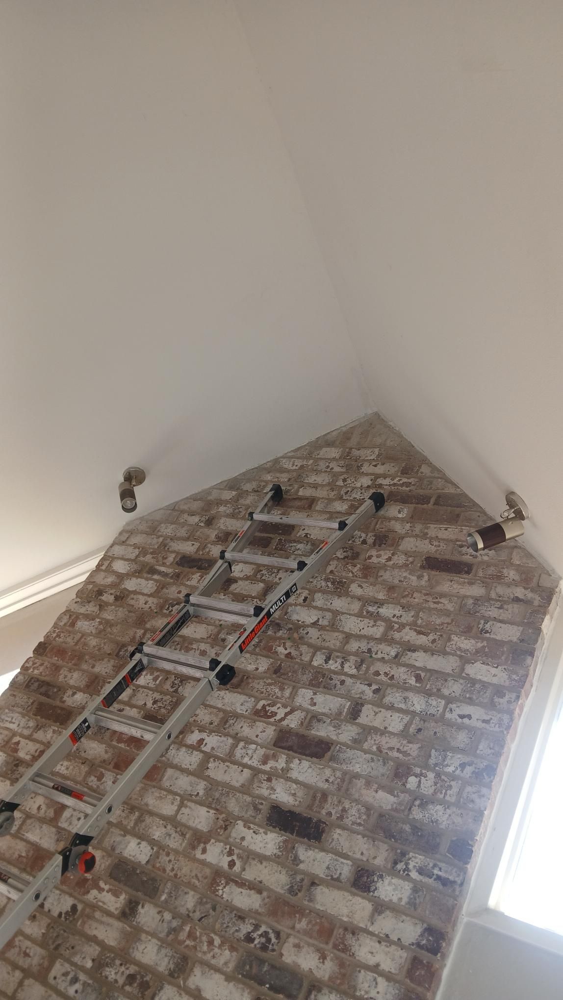 Ladder leaning against a brick wall beneath a white ceiling; two dark fixtures visible.