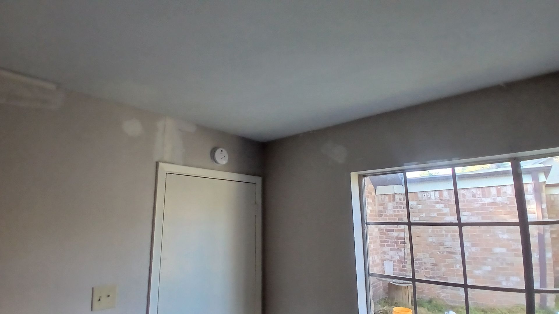 Interior room corner with white door, window, and smoke detector on the wall. Patched wall with neutral color paint.