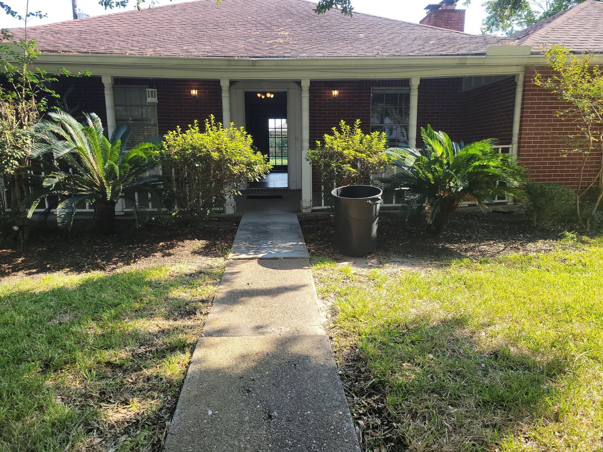 Brick house with porch, walkway to front door. Landscaping includes bushes and palm trees.